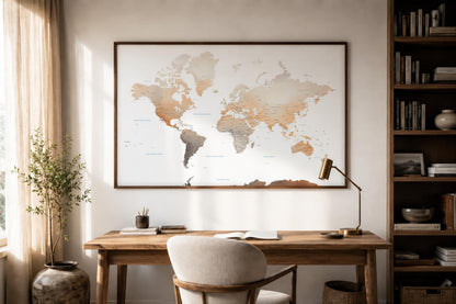 A large framed world map print hangs on a light beige wall above a wooden desk in a styled home office. The room includes a white upholstered chair, a brass desk lamp, a potted plant, and a tall bookshelf filled with books.