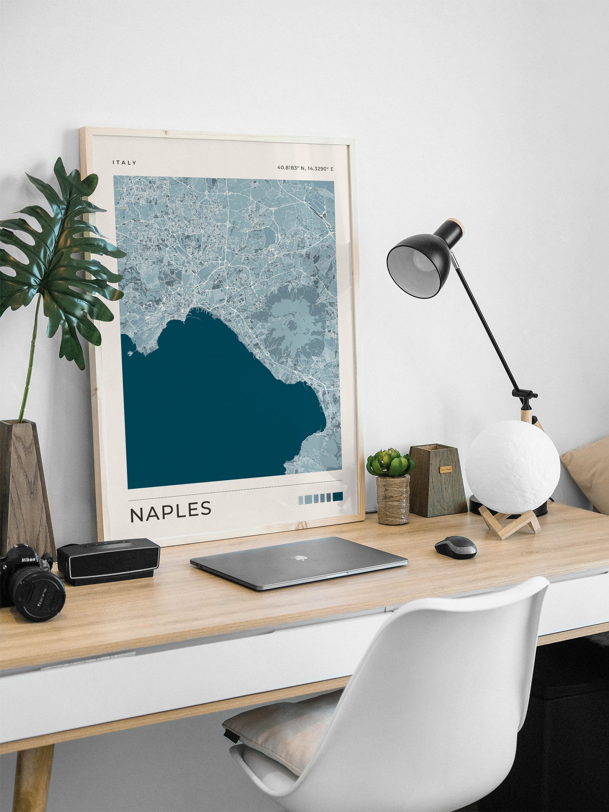 A framed Naples map print with a light wood frame leans on a light wood desk in a modern home office setup, alongside a laptop, camera, lamp, and white chair.