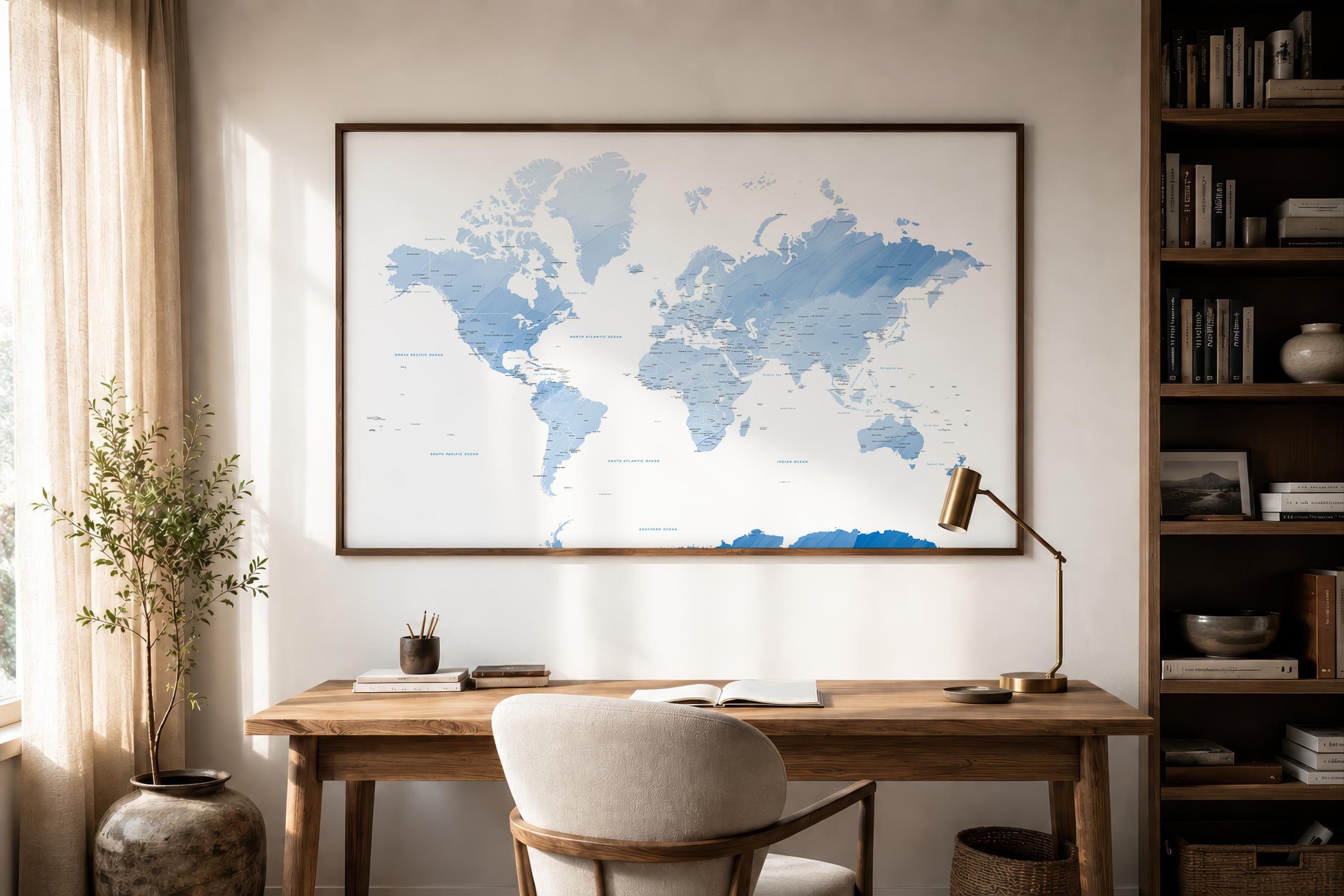 A large framed world map print in shades of blue hangs on a beige wall above a wooden desk in a styled home office with a chair, lamp, bookshelf, and potted plant.