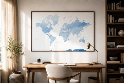 A large framed world map print in shades of blue hangs on a beige wall above a wooden desk in a styled home office with a chair, lamp, bookshelf, and potted plant.