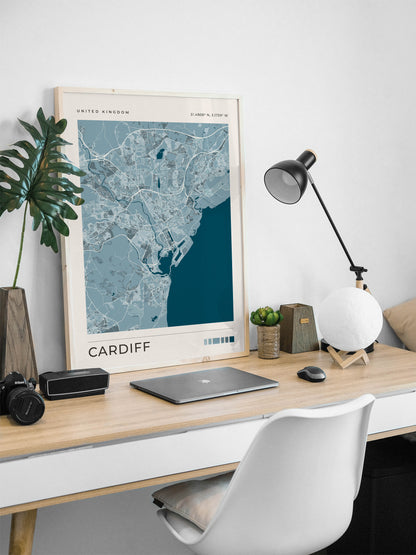 A framed Cardiff city map print leaning on a light wood desk in a modern home office setup, alongside a laptop, black desk lamp, white globe lamp, camera, and a white chair.