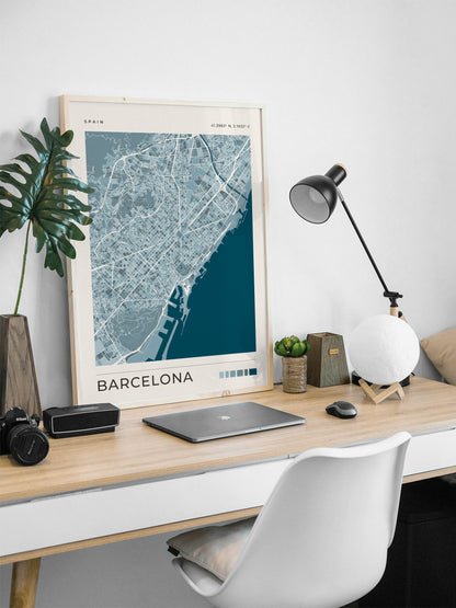 A framed Barcelona city map print leaning on a light wood desk in a modern home office setup with a laptop, lamp, plant, and white chair.