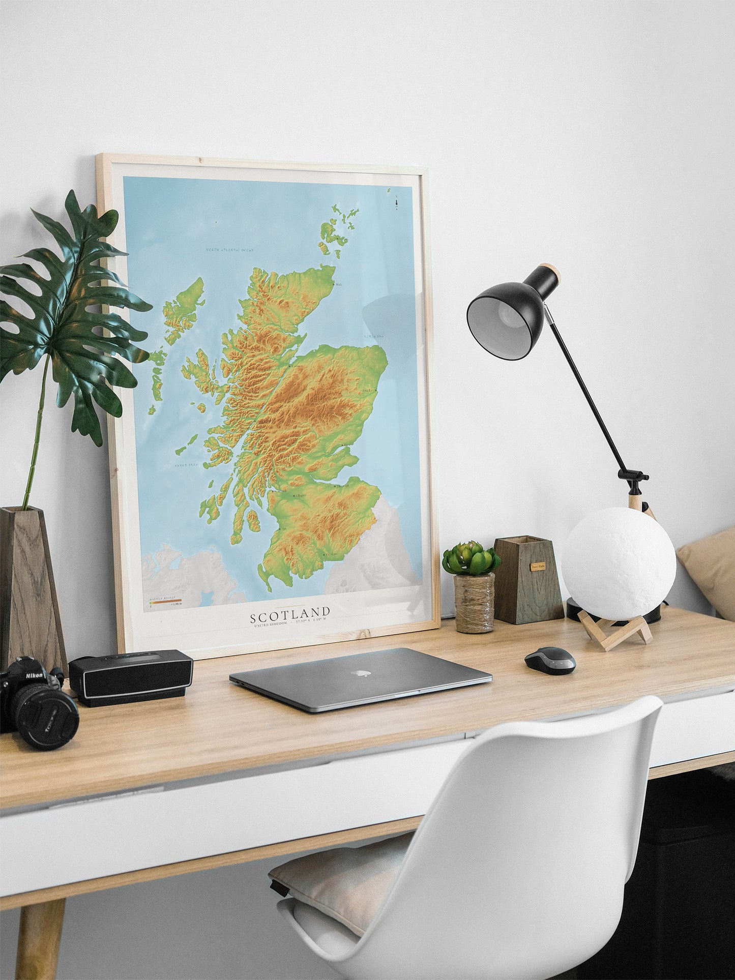 A framed topographical map of Scotland, featuring green and yellow terrain shading on a light blue background, styled on a light wood desk with a laptop, lamp, and plant.