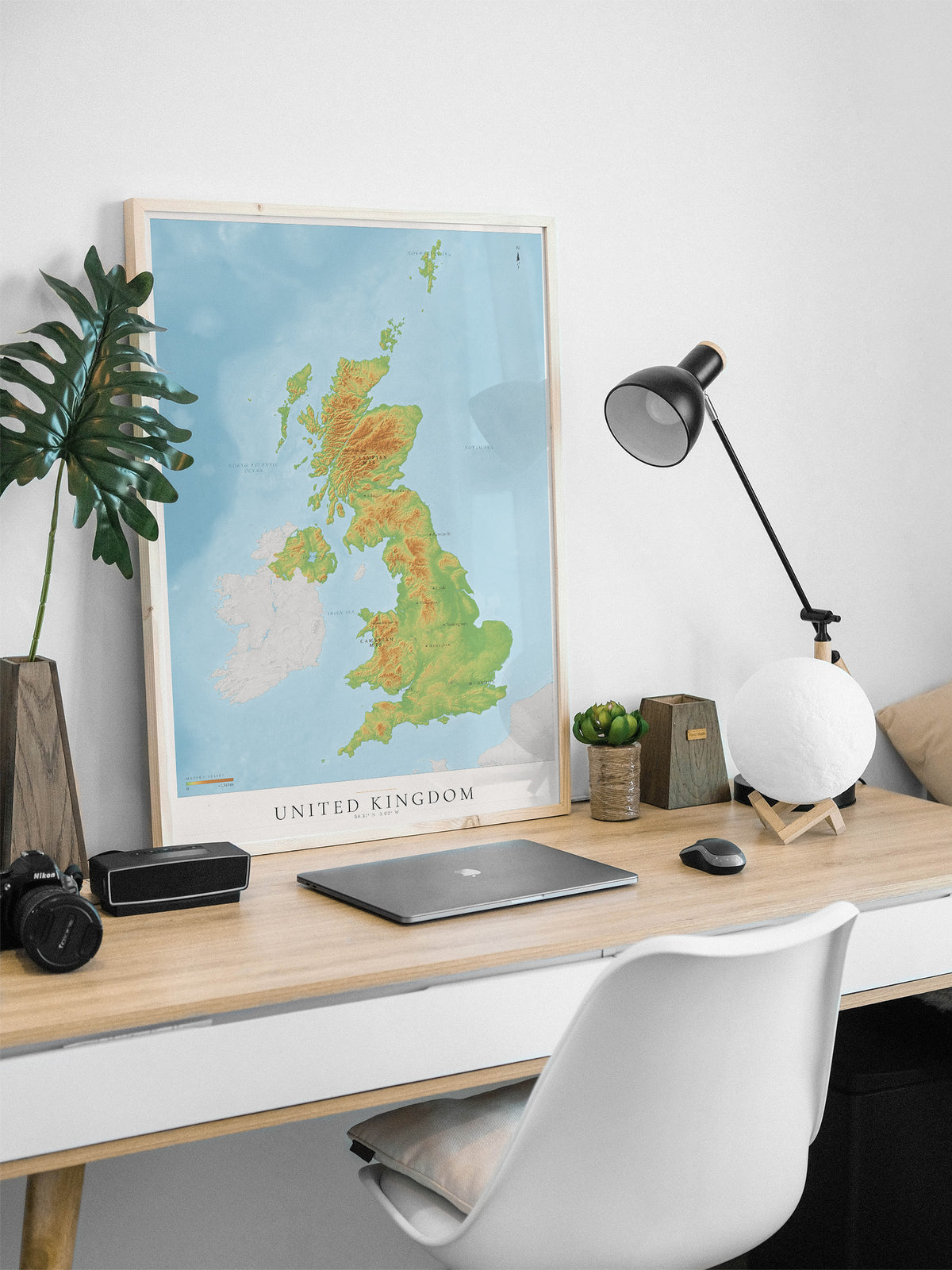 A framed vintage-style map of the United Kingdom leaning on a light wood desk in a modern home office setup, surrounded by a laptop, camera, lamp, and white chair against a white wall.
