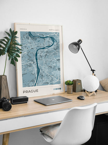 A framed art print of a Prague city map with a light blue and gray design, styled on a light wood desk with a laptop, lamp, and plant.