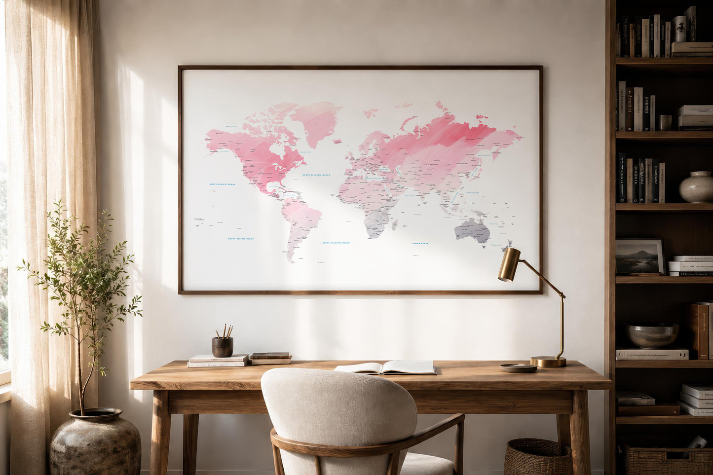 A large framed world map print with pink, coral, and gray watercolor-style continents hanging on a light wall above a wooden desk in a styled home office.