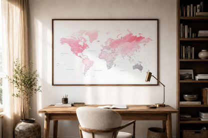 A large framed world map print with pink, coral, and gray watercolor-style continents hanging on a light wall above a wooden desk in a styled home office.
