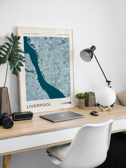 A framed art print of a blue and white street map of Liverpool, UK, displayed on a light wood desk in a modern home office setting with a white chair, black desk lamp, and green plant.