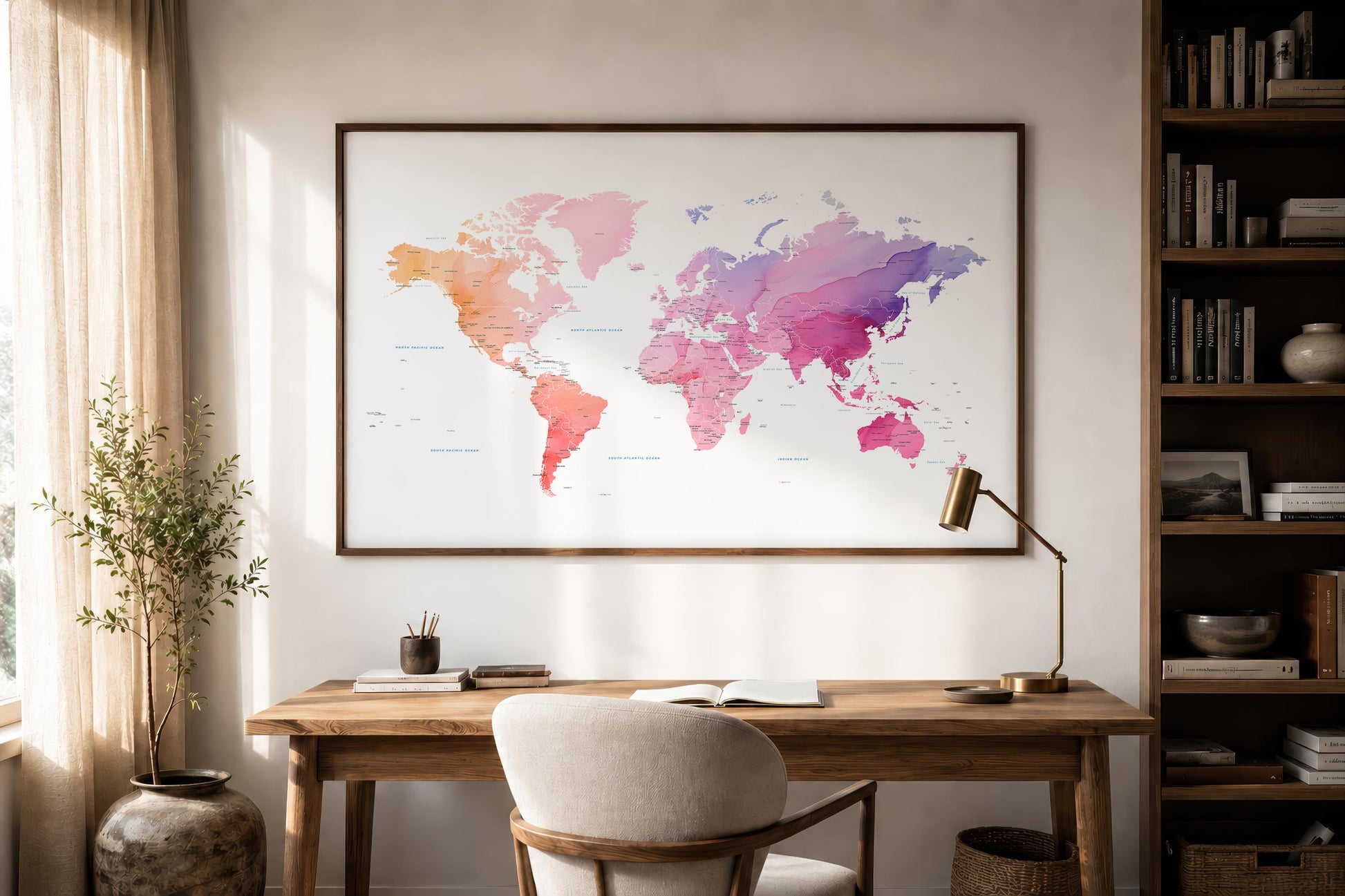 A framed world map print with a watercolor design in pink, purple, orange, and blue tones, displayed on a beige wall above a wooden desk in a styled home office setting.
