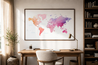 A framed world map print with a watercolor design in pink, purple, orange, and blue tones, displayed on a beige wall above a wooden desk in a styled home office setting.