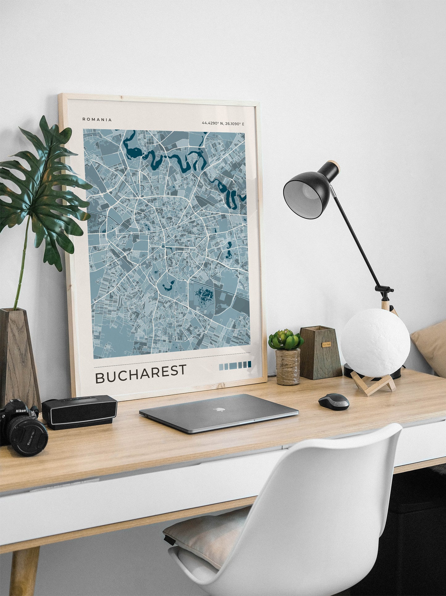 A framed blue map print of Bucharest, Romania, styled on a light wood desk with a laptop, lamp, and plant in a bright home office setting.