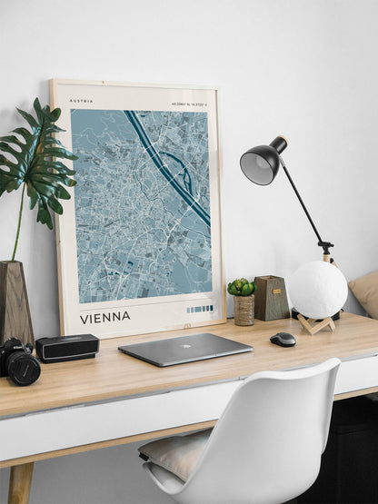 A framed Vienna city map print leaning on a light wood desk in a modern home office setup, alongside a laptop, black camera, plant, lamp, and white chair.
