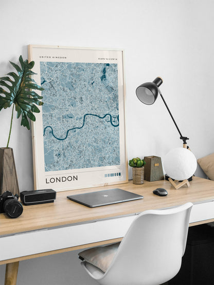 A framed blue-toned London city map print leaning on a light wood desk in a modern home office setup, with a laptop, lamp, plant, and white chair visible.