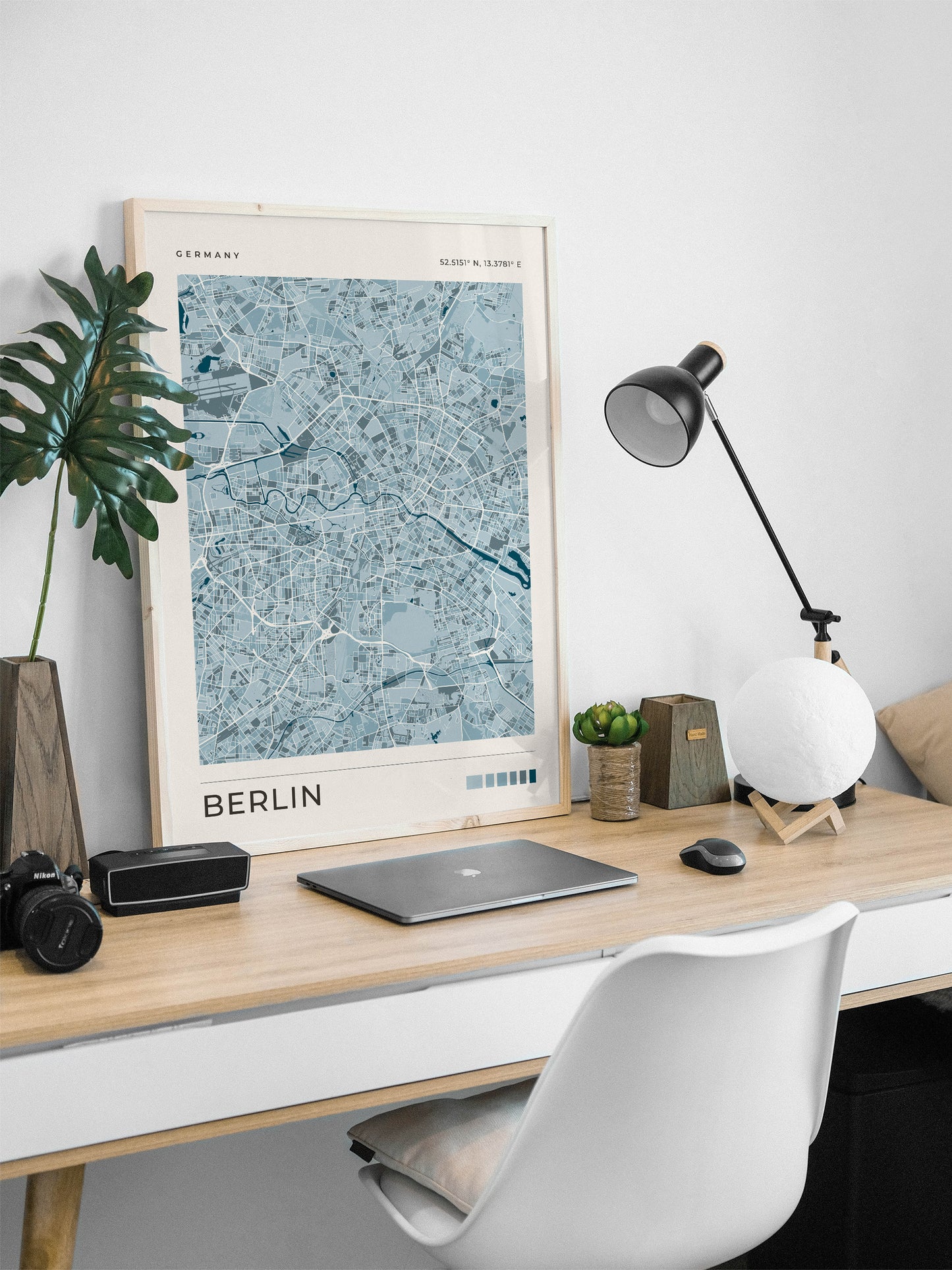 A framed blue map print of Berlin, Germany, styled on a light wood desk with a laptop, camera, lamp, and plant against a white wall.