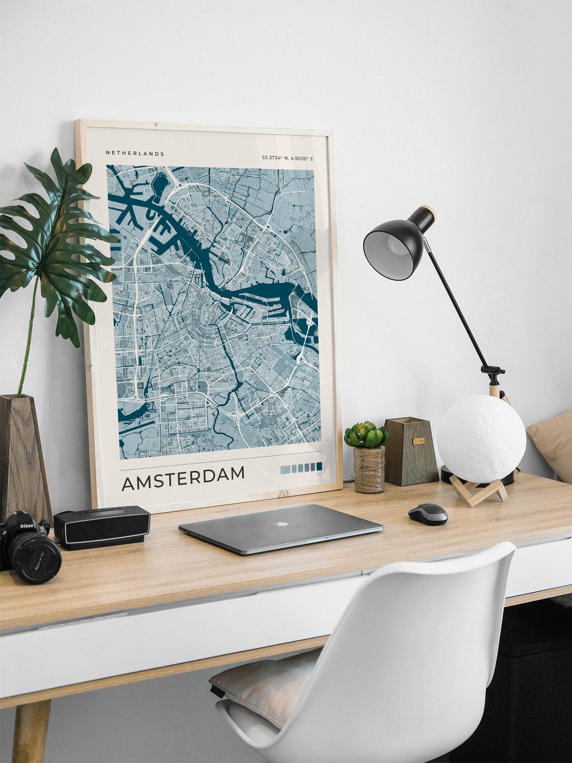 A framed Amsterdam city map print in blue and white hangs on a light wood desk in a modern home office setting, surrounded by a laptop, camera, lamp, and plant.