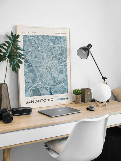 A framed San Antonio city map print with a light wood frame leans on a light wood desk in a modern home office setup, surrounded by a laptop, camera, lamp, and plant.