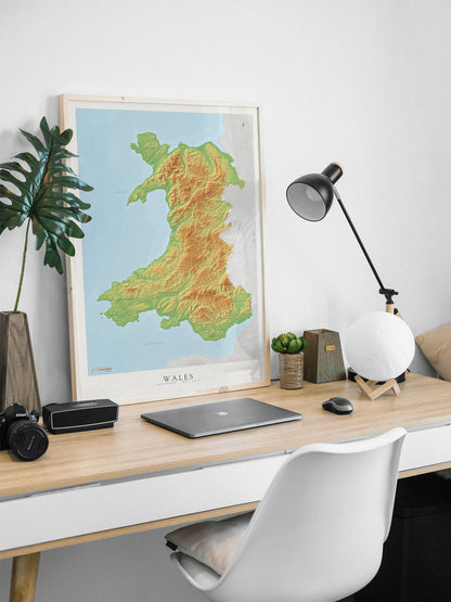 A framed topographical map of Wales leaning on a light wood desk in a modern home office setup, alongside a laptop, camera, lamp, and white chair.