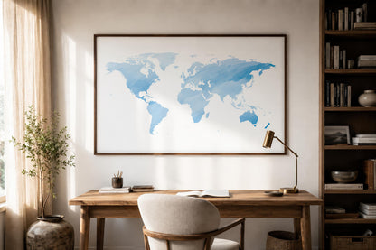 A large framed world map print with watercolor-style blue continents hangs on a beige wall above a wooden desk in a styled home office setting with a chair, lamp, and bookshelves.