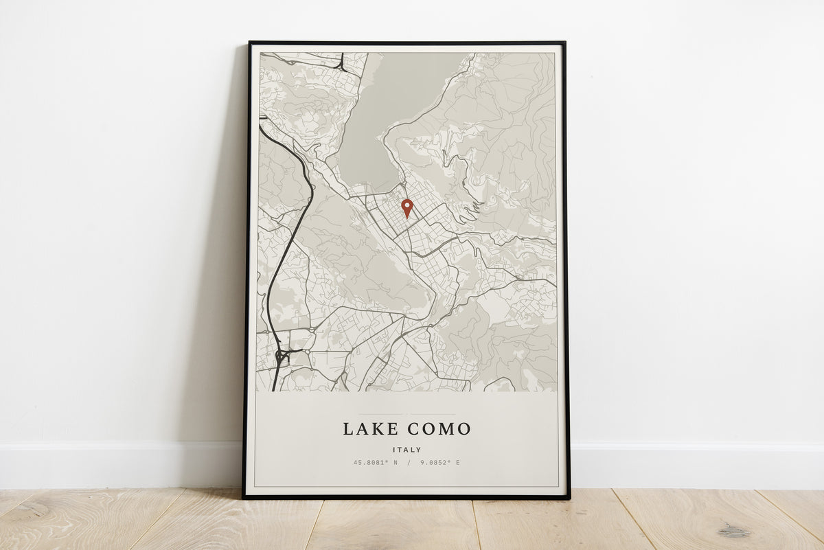 A framed minimalist map print of Lake Como, Italy, leaning against a white wall on a light wood floor. The print features a light beige and gray map with a small red pin marker and the text 'LAKE COMO ITALY' at the bottom.