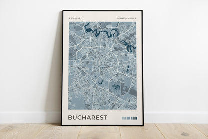 A framed minimalist city map print of Bucharest, Romania, leaning against a white wall on a light wood floor. The print features a light blue and gray street map with dark blue river lines on a cream background, enclosed in a thin black frame.