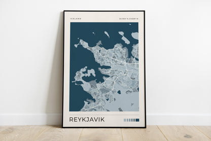 A framed minimalist map print of Reykjavík, Iceland, leaning against a white wall on a light wood floor. The print features a dark teal and light gray map design with 'REYKJAVIK' printed at the bottom.