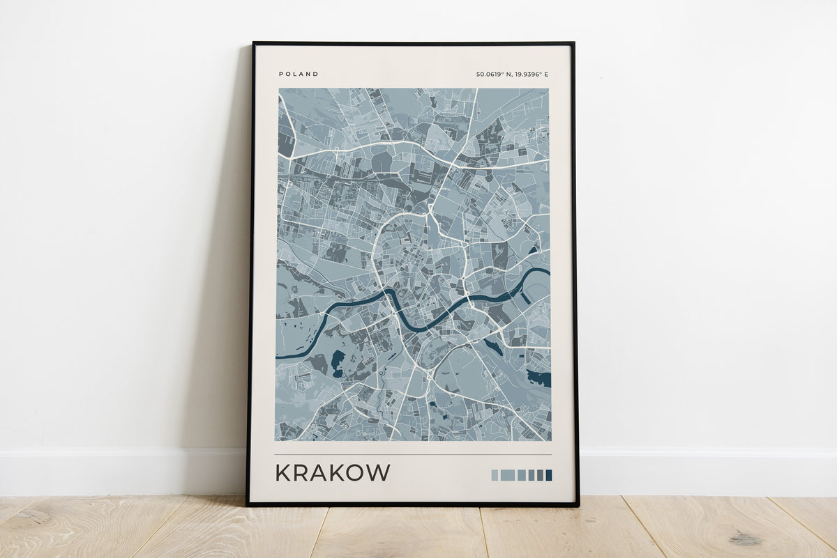 A framed minimalist map poster of Krakow, Poland, leaning against a white wall on a light wood floor. The poster features a light blue and gray street grid with a dark blue river, set in a thin black frame.