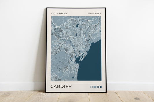 A framed minimalist map print of Cardiff, United Kingdom, leaning against a white wall on a light wood floor. The print features a muted blue and gray street map with a dark teal ocean section, enclosed in a thin black frame.