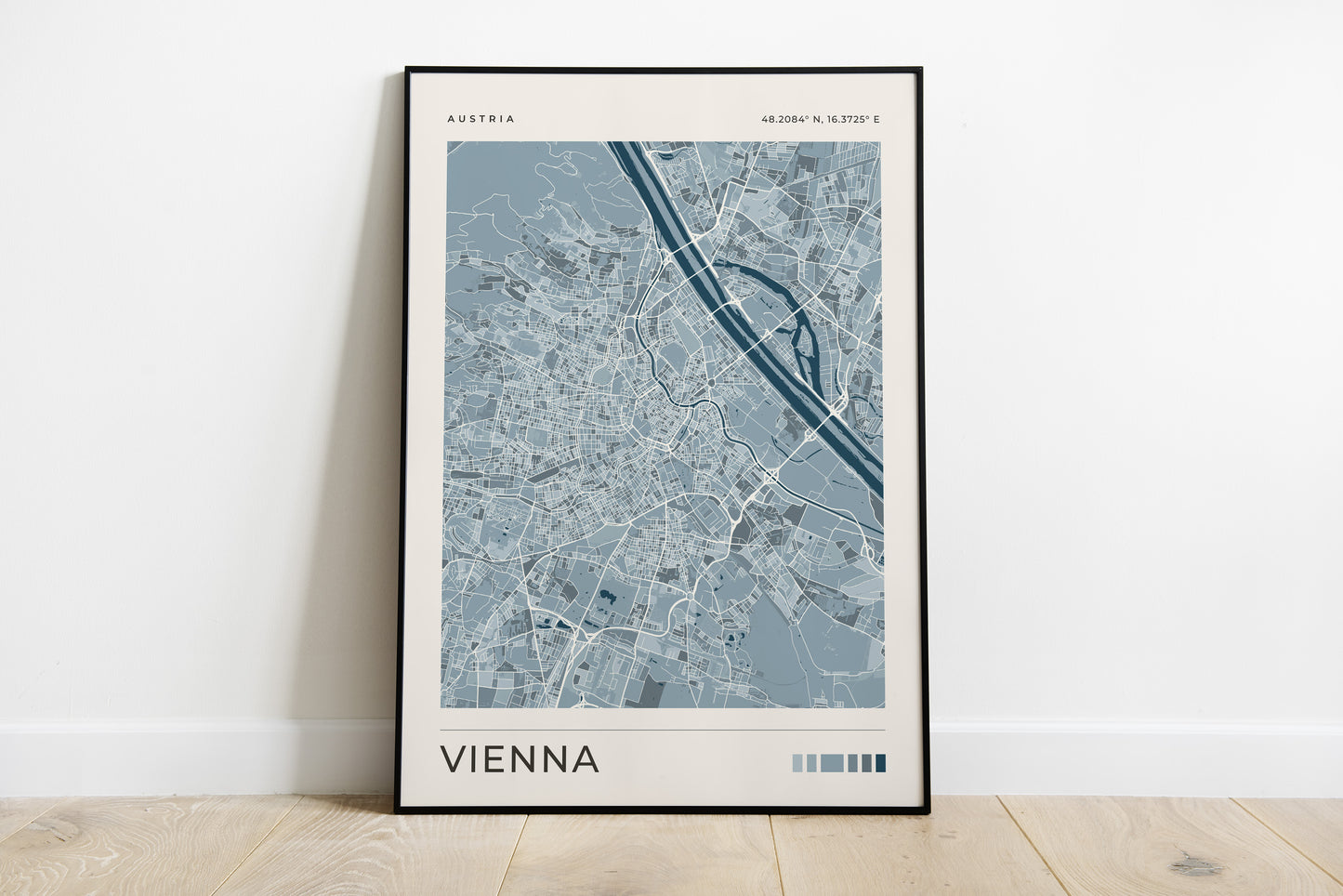 A framed minimalist city map print of Vienna, Austria, leaning against a white wall on a light wood floor.