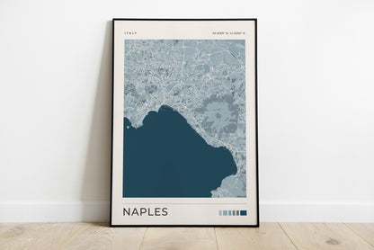 A framed map art print of Naples, Italy, leaning against a white wall on a light wood floor. The print features a modern map design in shades of blue and gray with the city name 'NAPLES' printed at the bottom.