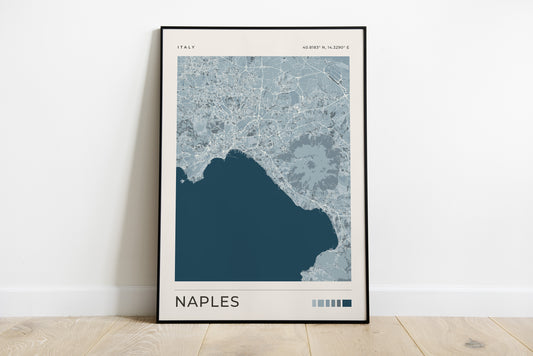 A framed map art print of Naples, Italy, leaning against a white wall on a light wood floor. The print features a modern map design in shades of blue and gray with the city name 'NAPLES' printed at the bottom.
