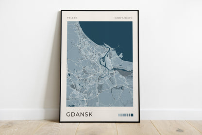 A framed city map print of Gdansk, Poland, leaning against a white wall on a light wood floor. The print features a modern map design in shades of blue, gray, and white, with the city name 'GDANSK' printed at the bottom.