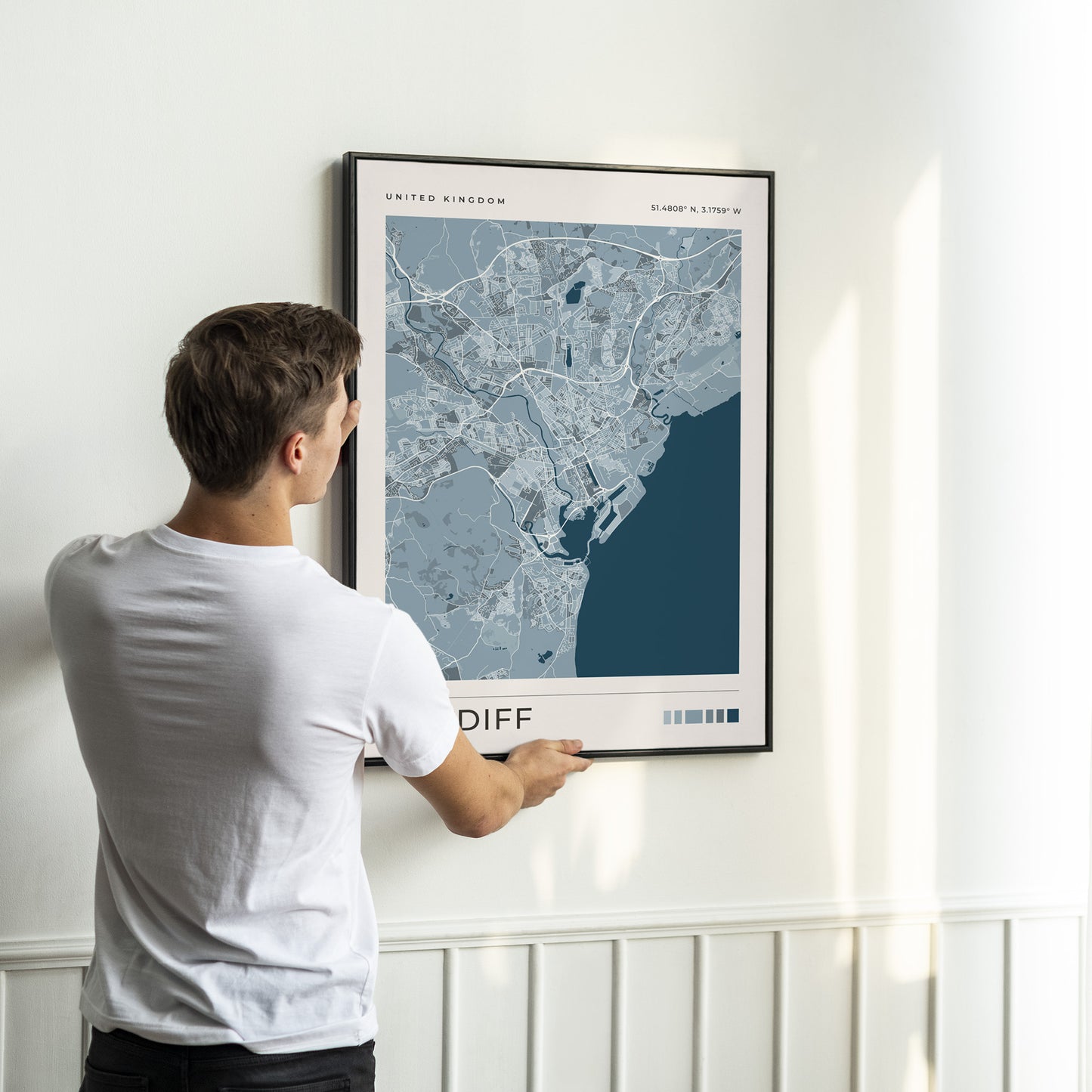 A man in a white t-shirt is shown from behind, holding a framed map print of Cardiff, United Kingdom, against a white wall with wainscoting.