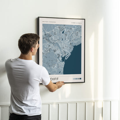 A man in a white t-shirt is shown from behind, holding a framed map print of Cardiff, United Kingdom, against a white wall with wainscoting.
