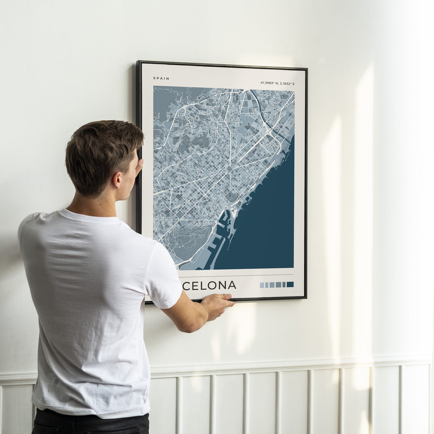 A man in a white t-shirt is shown from behind, adjusting a framed map print of Barcelona on a white wall.