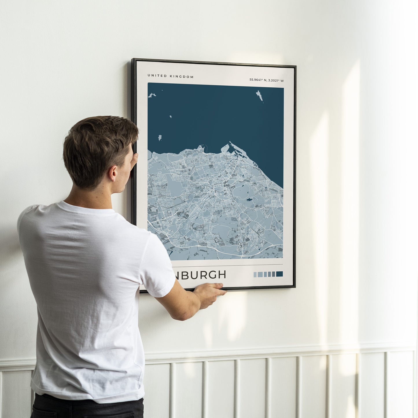 A man in a white t-shirt is shown from behind, holding a framed Edinburgh map print against a white wall with wainscoting.