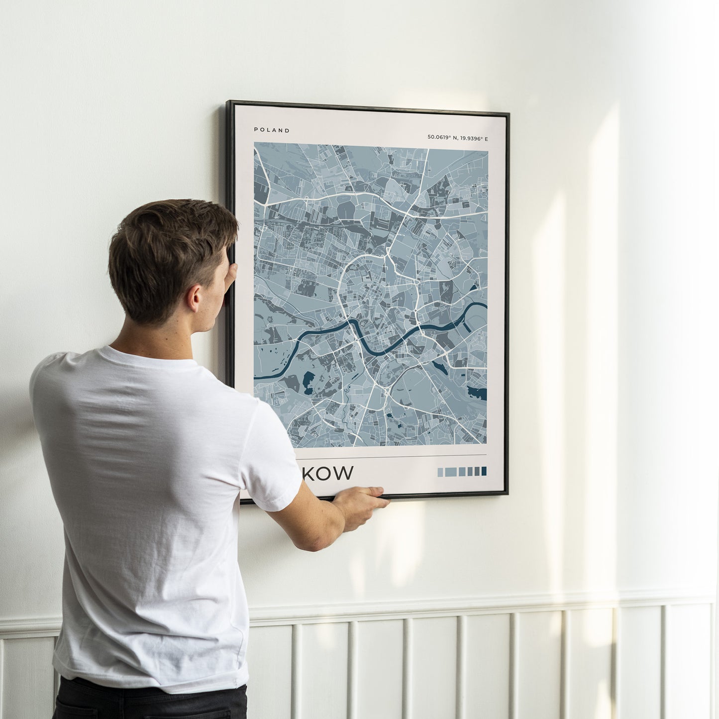 A man in a white t-shirt is shown from behind, adjusting a framed city map print of Warsaw, Poland, on a white paneled wall.