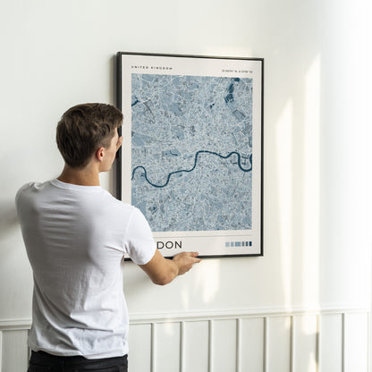 A man in a white t-shirt is shown from behind, holding a framed London city map print against a white wall.