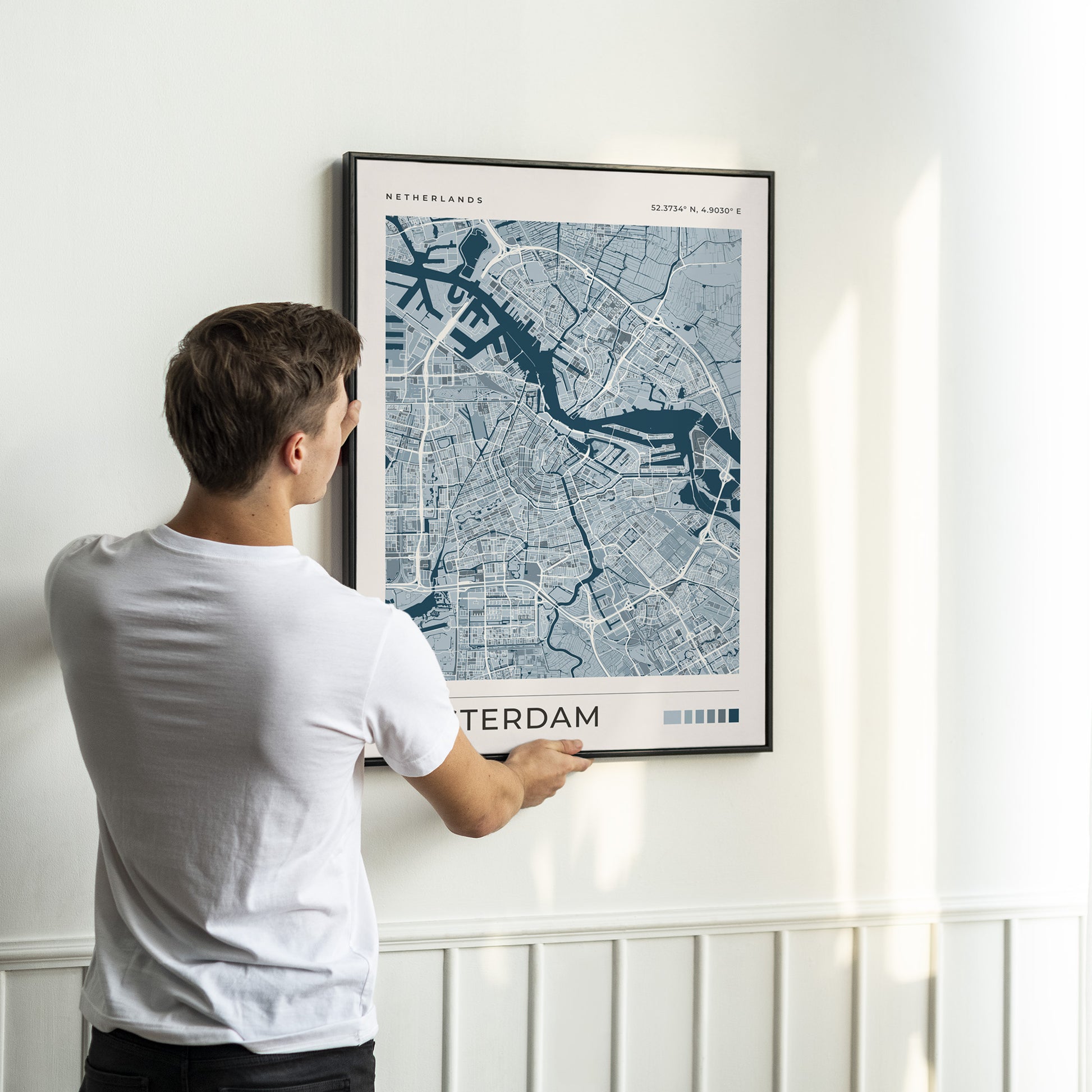 A man in a white t-shirt is shown from behind, holding a framed city map poster of Amsterdam, Netherlands, against a white wall with wainscoting.