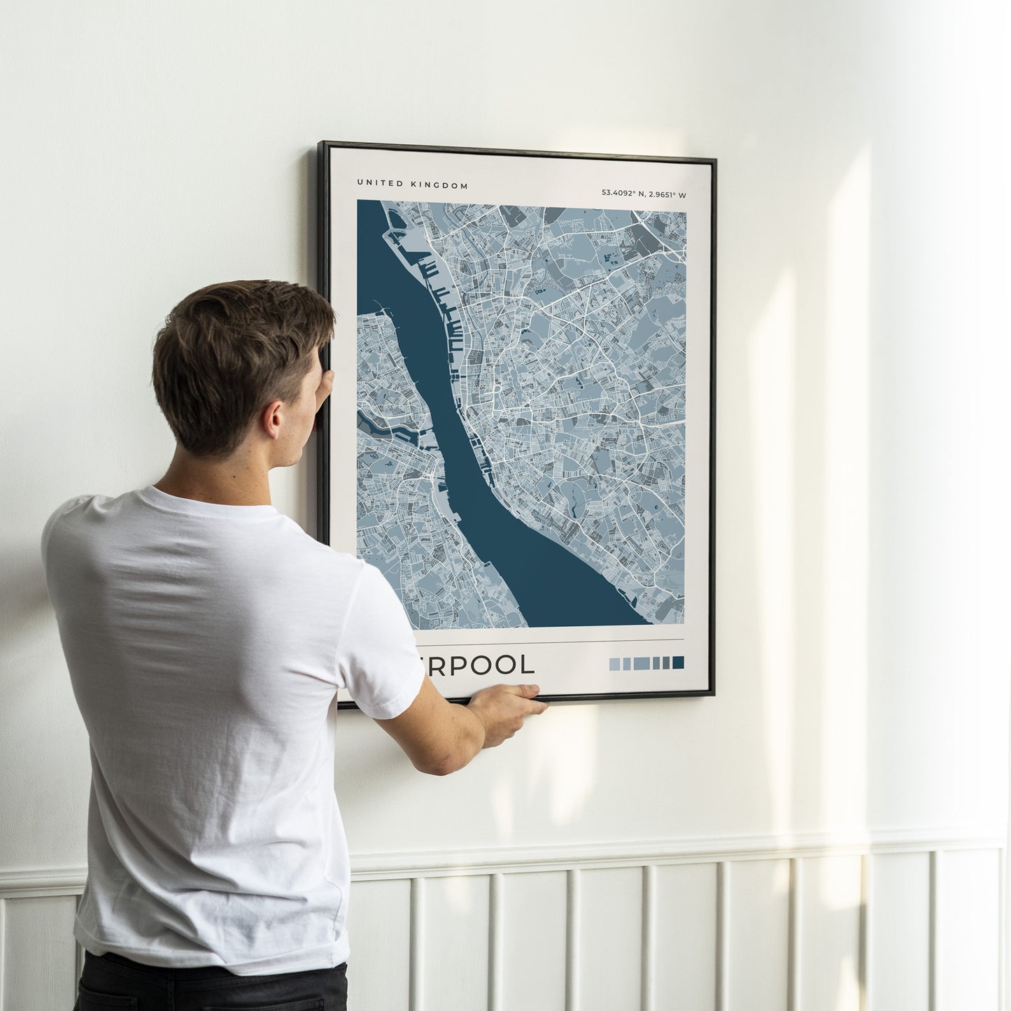 A man in a white t-shirt is shown from behind, holding a framed map print of Liverpool, United Kingdom, against a white wall with sunlight streaming in.