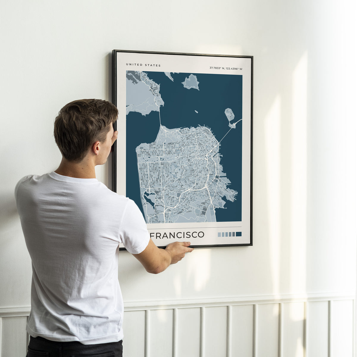 A man in a white t-shirt is shown from behind, holding a framed print of a stylized map of San Francisco, California. The print features a dark navy blue background with light gray city details and the text 'UNITED STATES' and 'SAN FRANCISCO'.