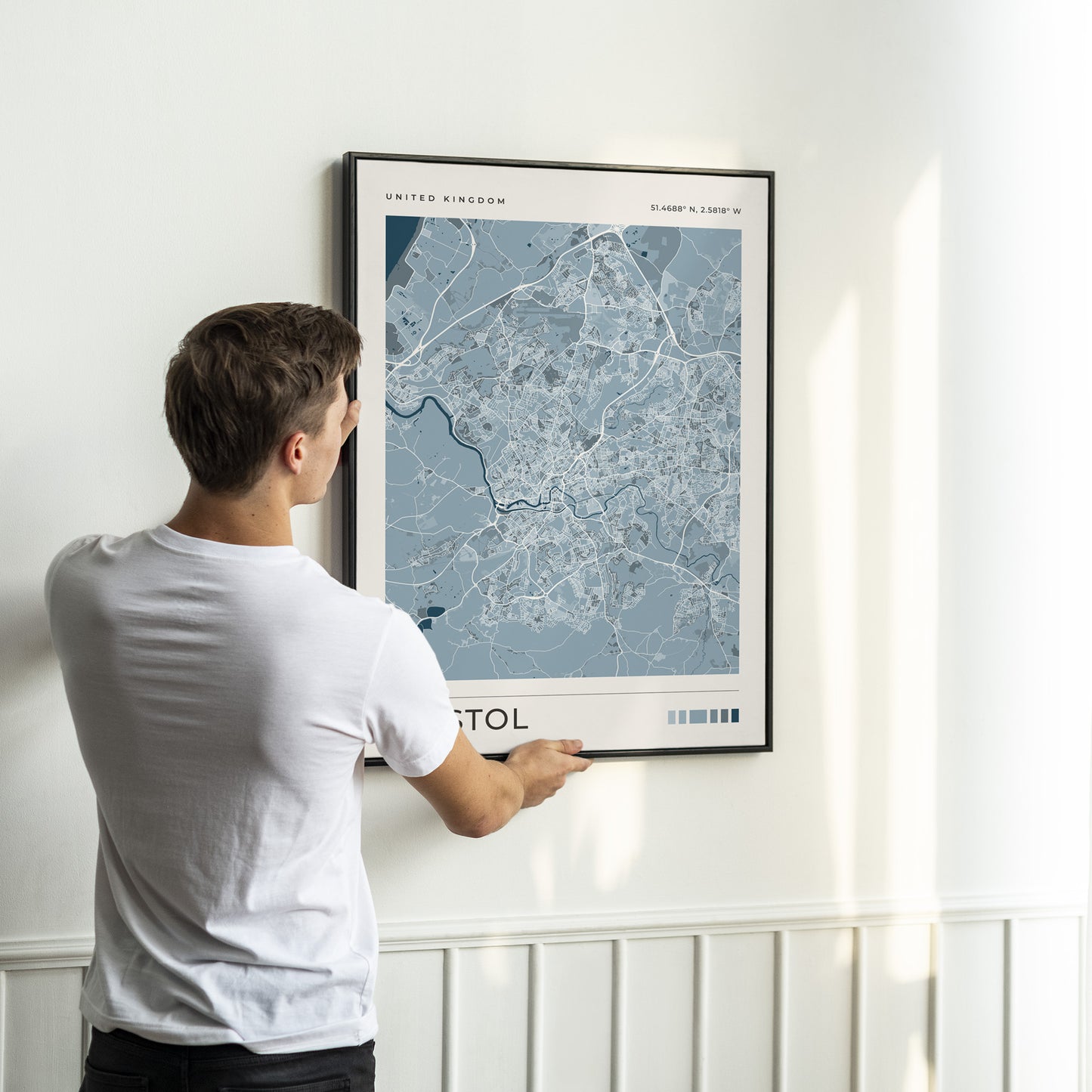 A man in a white t-shirt is shown from behind, holding a framed map print of London, United Kingdom, against a white wall with wainscoting.