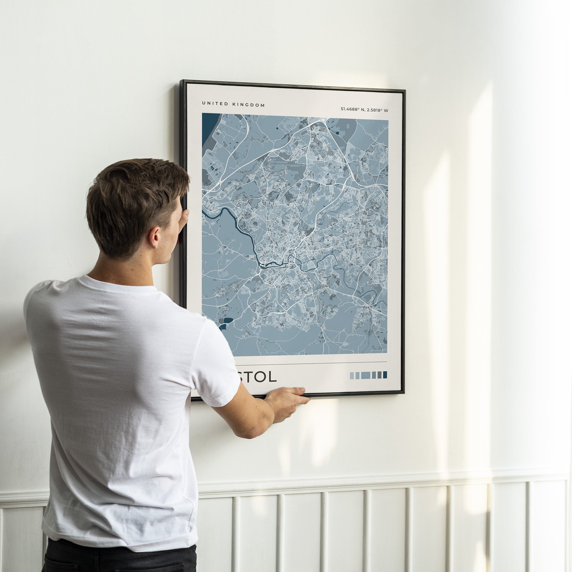 A man in a white t-shirt is shown from behind, holding a framed map print of London, United Kingdom, against a white wall with wainscoting.