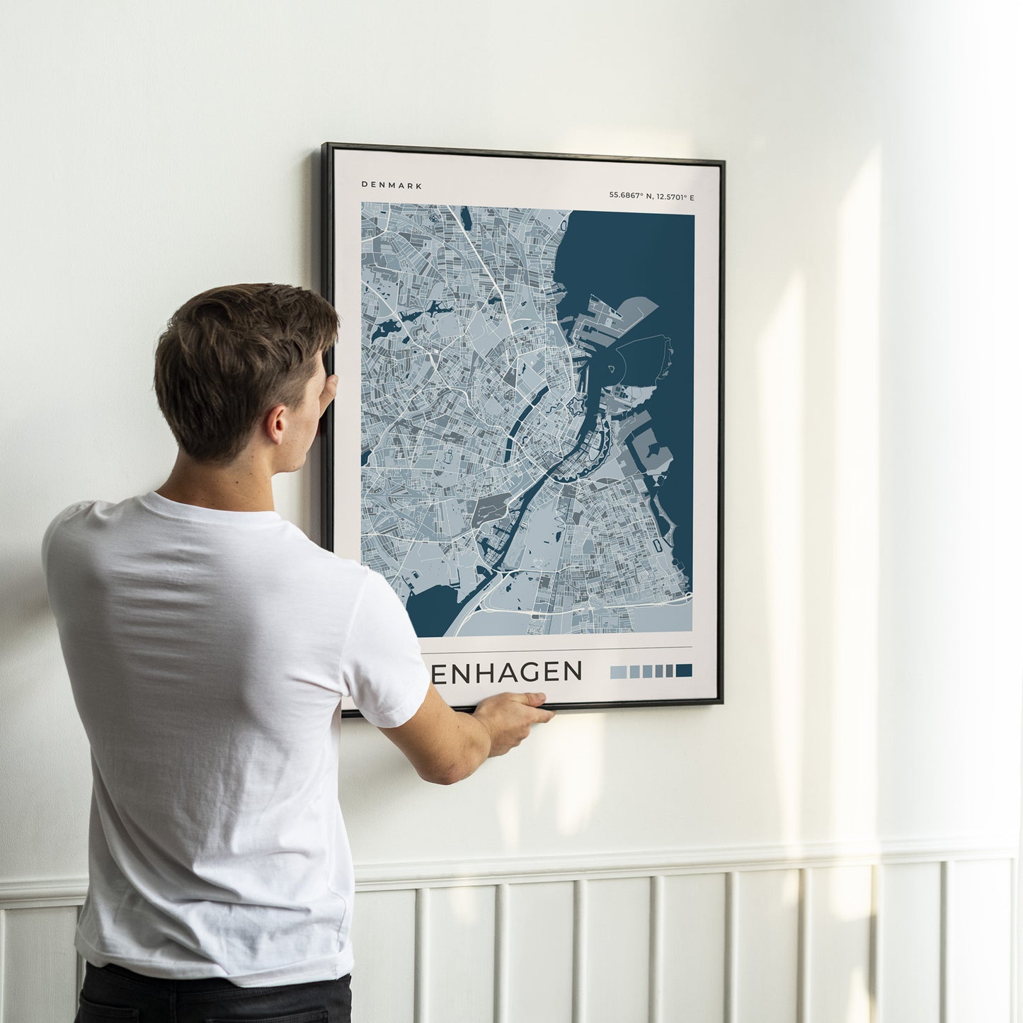 A man in a white t-shirt is shown from behind, holding a framed map print of Copenhagen, Denmark. The print features a modern blue and gray street map design with the word 'COPENHAGEN' at the bottom, mounted on a white wall with a wainscoted baseboard.