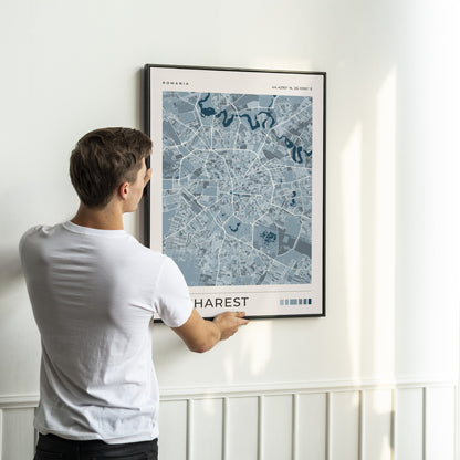 A man in a white t-shirt is shown from behind, holding a framed map print of Harest, Romania, on a white wall with wainscoting.