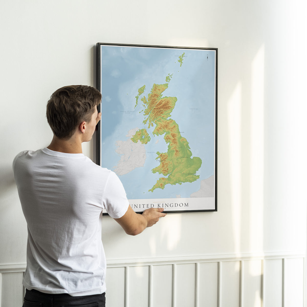 A man in a white t-shirt is shown from behind, holding a framed topographical map of the United Kingdom on a white wall.
