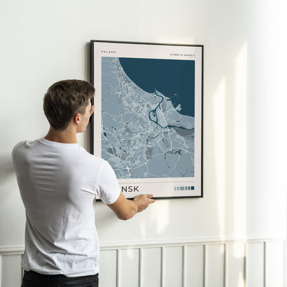 A man in a white t-shirt is shown from behind, holding a framed city map print of Oslo, Norway. The print features a modern design with a dark blue sea and light blue-gray land, framed in black and hung on a white wall with wainscoting below.