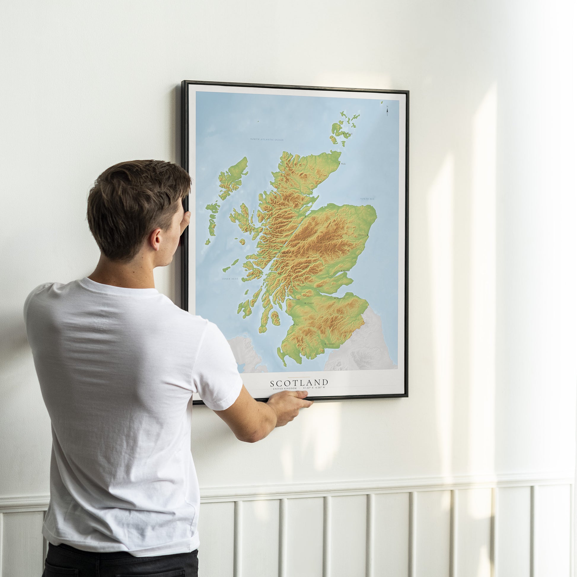A man in a white t-shirt is shown from behind, holding a framed topographical map of Scotland on a white wall.