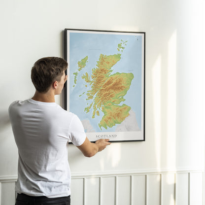 A man in a white t-shirt is shown from behind, holding a framed topographical map of Scotland on a white wall.
