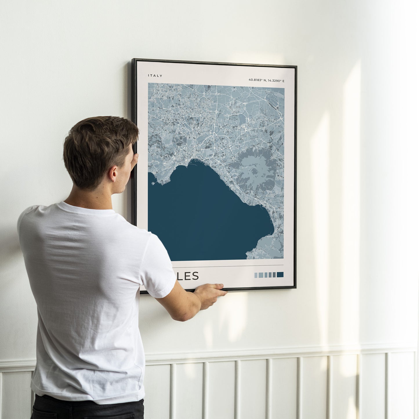 A man in a white t-shirt is shown from behind, holding a framed map print of Naples, Italy, against a white wall with wainscoting.