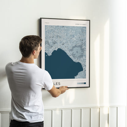 A man in a white t-shirt is shown from behind, holding a framed map print of Naples, Italy, against a white wall with wainscoting.